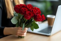 Woman holding fresh red rose bouquet while working at laptop, romantic lifestyle moment captured in soft natural lighting at wooden desk.
