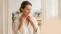 Young Caucasian woman in white robe touching neck with gentle smile, eyes closed in peaceful moment. Morning skincare routine and self-care wellness concept.