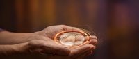 Elderly hands gently holding glowing compass with warm light emanating from palms against blurred background suggesting guidance and direction.