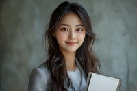 Young Asian businesswoman with long dark wavy hair and natural makeup holding notebook, wearing grey blazer, smiling warmly against textured wall background.