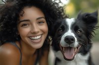 Young African American woman with natural curly hair smiling next to happy Border Collie dog outdoors, close-up portrait showing genuine joy and connection between pet and owner.