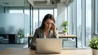 Stressed businesswoman with headache working on laptop in modern office. Professional female executive experiencing work pressure and fatigue at workplace.