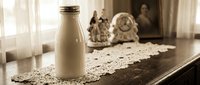 Fresh milk bottle on vintage lace doily creates rustic farmhouse kitchen scene with warm natural lighting and traditional home decor elements.