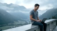 Young man working on laptop outdoors in mountain valley with misty peaks. Remote work lifestyle and digital nomad concept for modern business.