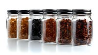 Glass jars filled with various loose leaf tea varieties arranged in row showing different colors and textures from golden to black on white background with soft reflection.