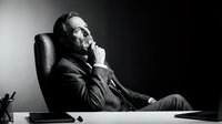 Mature man with beard in business suit sitting at desk with hand to face, contemplative expression, black and white portrait for corporate and editorial use.