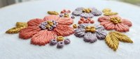 Hand embroidered floral design with coral and purple flowers on white fabric featuring detailed stitching and golden leaves for craft projects.