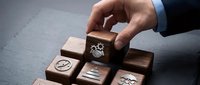 Businessman hand selecting wooden cube with gear mechanism symbol representing process optimization and workflow management in corporate environment.