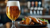 Cold glass of light beer with bubbles and foam head next to plate of spicy chicken wings at bar counter, selective focus on beverage.