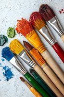 Paint brushes with colorful acrylic paint smears arranged on white textured background. Wooden handles and silver ferrules show artistic tools in vibrant blue, yellow, orange and green.