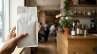 Cropped hand holding translucent reusable shopping bag in modern cafe interior with blurred counter background. Sustainable lifestyle and eco-friendly retail concept.