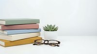 Stack of colorful books with eyeglasses and small succulent plant on white desk. Minimalist workspace setup for reading, studying and education concepts.