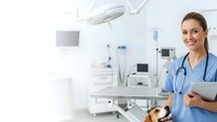 Smiling female doctor in blue scrubs holding tablet in modern hospital operating room with medical equipment and surgical lights.