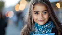 Young girl with long brown hair wearing blue patterned scarf smiling at camera against blurred bokeh background, conveying happiness and innocence.