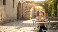 Young blonde boy in striped shirt sitting at outdoor cafe table in charming European cobblestone street with warm evening lights and historic stone architecture.