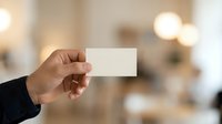 Hand holding blank white business card in front of blurred office background with warm lighting, suitable for branding and corporate identity mockups.