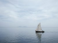 Lone sailboat gliding on calm sea under overcast sky, distant mountains on horizon, peaceful nautical scene evoking freedom and solitude.