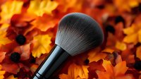 Professional makeup brush with soft synthetic bristles lying on vibrant orange and red autumn maple leaves creating seasonal beauty composition.