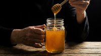 Golden honey dripping from wooden dipper into glass jar held by hands on rustic wooden table against dark background for natural food photography.