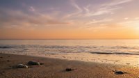 Serene sandy beach at golden hour with gentle waves rolling onto shore, scattered rocks in foreground, and soft pastel sky reflecting warm sunset light over calm ocean waters.