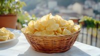 Crispy potato chips piled high in rustic wicker basket on white tablecloth outdoors, natural sunlight creates appetizing glow on golden snacks.