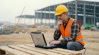 Construction worker in safety gear using laptop computer at building site for project management and digital planning tasks.