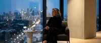Professional businesswoman in black suit sitting in modern office chair overlooking illuminated city skyline at dusk through floor-to-ceiling windows.
