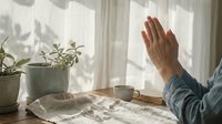 Person practicing morning meditation with raised hand in sunlit bedroom with houseplants creating peaceful mindful wellness routine for mental health and relaxation.