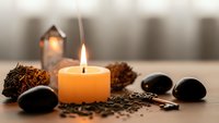 Relaxing spa scene with burning candle, black stones, pine cones and lavender seeds creating peaceful wellness atmosphere for meditation and aromatherapy treatments.