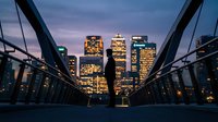 Urban skyline at dusk with illuminated office buildings viewed through modern bridge architecture creating dramatic cityscape perspective for business concepts.