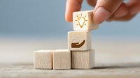 Hand placing wooden cube with light bulb icon above hand symbol, representing idea support and innovation concept on stacked blocks.