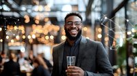 Young African American businessman in glasses and blazer smiling while holding champagne at corporate party with festive bokeh lights and blurred background.