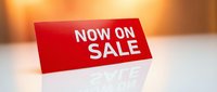 Red NOW ON SALE sign standing on reflective white surface with warm bokeh background, promoting retail discount and special offer event.
