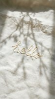 Word hello embroidered in cream thread on soft linen fabric with natural branch shadows, warm sunlight, cozy aesthetic mood.