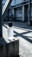 Minimalist white cosmetic bottle with black cap on concrete block in industrial warehouse setting with dramatic shadows and metal architecture.