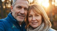 Happy mature Caucasian couple taking selfie outdoors during golden hour sunset, smiling at camera with warm natural lighting and bokeh background.