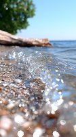 Crystal clear wave splashing on rocky beach shore with sparkling water drops and sunlight bokeh, peaceful summer seascape against blue sky background.