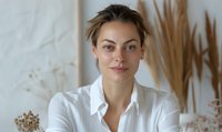 Young Caucasian woman with natural makeup and pulled back hair wearing white shirt sitting at table with dried pampas grass and floral decorations in light minimal interior.