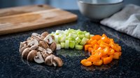 Fresh diced vegetables including mushrooms, celery and carrots arranged on dark cutting board for healthy cooking and meal preparation in modern kitchen.