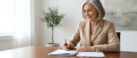 Professional middle-aged Caucasian businesswoman in beige blazer writing documents at modern office desk with plant, smiling confidently in bright workspace.