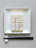 Fresh tofu cubes arranged in geometric pattern on white square plate with black chopsticks, minimalist Asian cuisine presentation on light gray background.