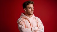 Young Caucasian male model wearing pink hoodie standing with crossed arms against red background, looking away with confident expression. Professional studio portrait.