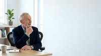 Senior businessman in dark blazer sitting at desk, hand to chin in thoughtful pose, contemplating business strategy or decision-making in modern office environment.