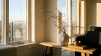 Warm sunlight streams through large windows illuminating minimalist interior with ceramic vase, dried branches and wooden furniture creating cozy atmosphere.