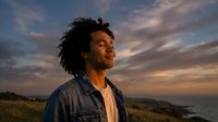 Young African American man with curly hair looking up thoughtfully at golden hour sunset sky, wearing denim jacket in peaceful outdoor setting.