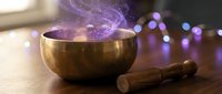 Tibetan singing bowl with purple mystical smoke and wooden mallet on table with bokeh lights creating spiritual meditation atmosphere for wellness practices.