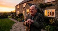 Elderly Caucasian man with walking stick standing in front of stone cottage during golden hour sunset, peaceful countryside retirement lifestyle.