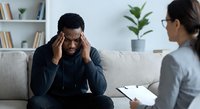 Stressed African American man holding head during therapy session with female counselor in modern office setting for mental health support.