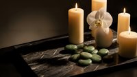Spa wellness setup with lit candles, green stones, and white orchid on dark marble tray, evoking relaxation and luxury treatment.