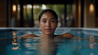Young Asian woman relaxing in indoor swimming pool with calm expression, warm evening lighting creates peaceful atmosphere for wellness and spa concept.